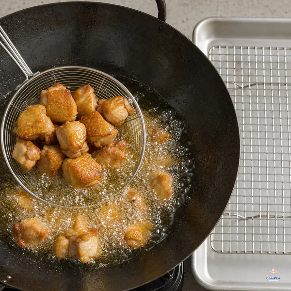 A wire skimmer lifts deep-golden skin-on chicken pieces from vigorously bubbling oil beside a draining rack