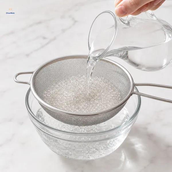 Cold water from a plain glass pitcher rinses hundreds of tiny transparent tapioca pearls in a fine sieve over a glass bowl