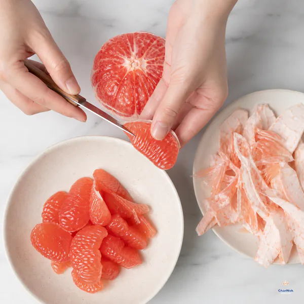 Hands cut a peeled pink grapefruit segment away from its membrane over a bowl of intact juice-filled fruit sacs