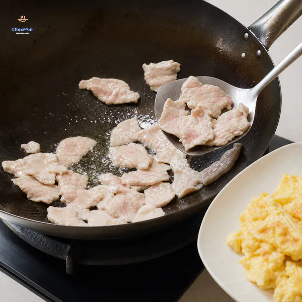 Separated thin pork slices have just turned pale with faint pink centres as a wok spatula lifts them toward a plate of glossy egg curds