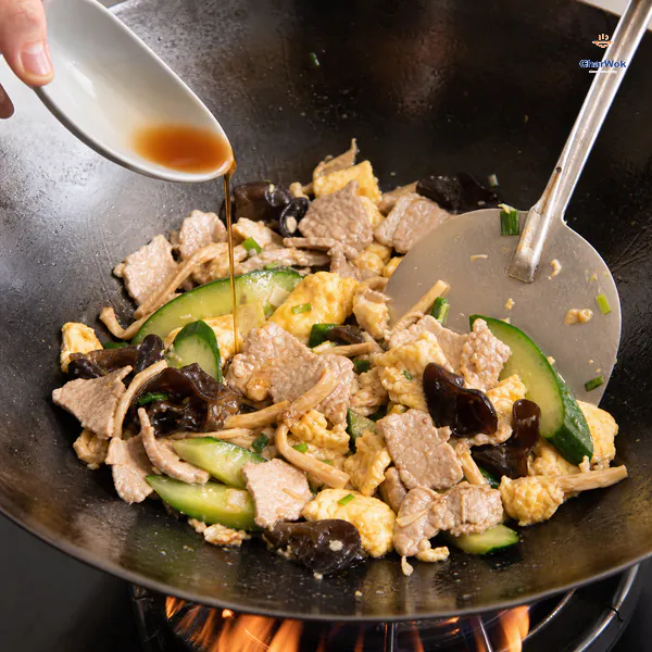 A small stream of light soy sauce falls over tender pork, glossy egg, wood ear, lily buds and bright cucumber during a quick wok toss