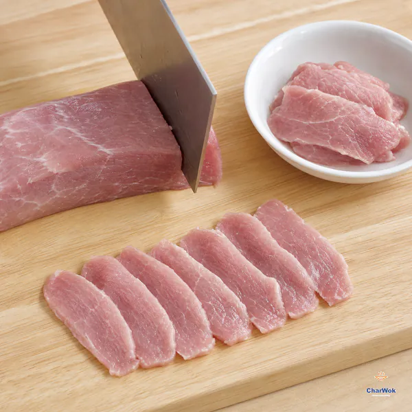 A cleaver cuts lean pork tenderloin across the grain while eight even thin slices and a bowl of sliced pork remain fully visible