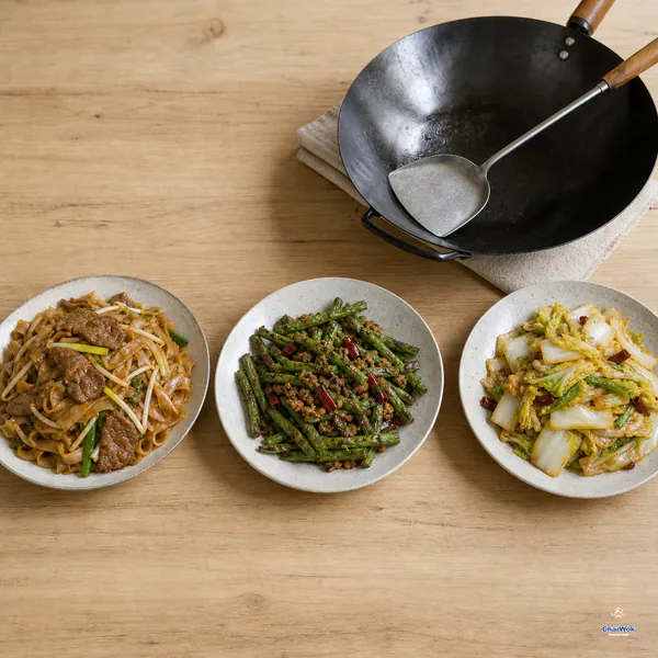 Dry beef chow fun, wrinkled dry-fried green beans and glossy hot-and-sour napa cabbage sit in three separate plates beside a carbon-steel wok