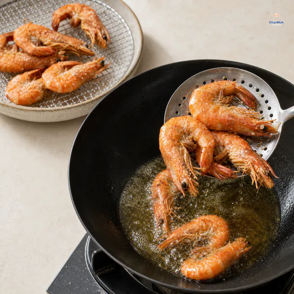 A perforated ladle lifts whole orange shell-on shrimp from bubbling oil toward a wire rack holding a dry crisp batch