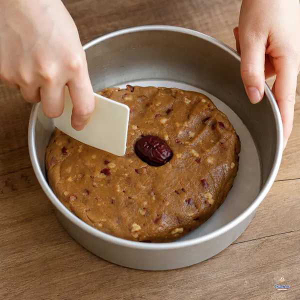 A spatula presses firm brown jujube-walnut dough flat in an oiled round tin lined with parchment, with one whole date at the centre