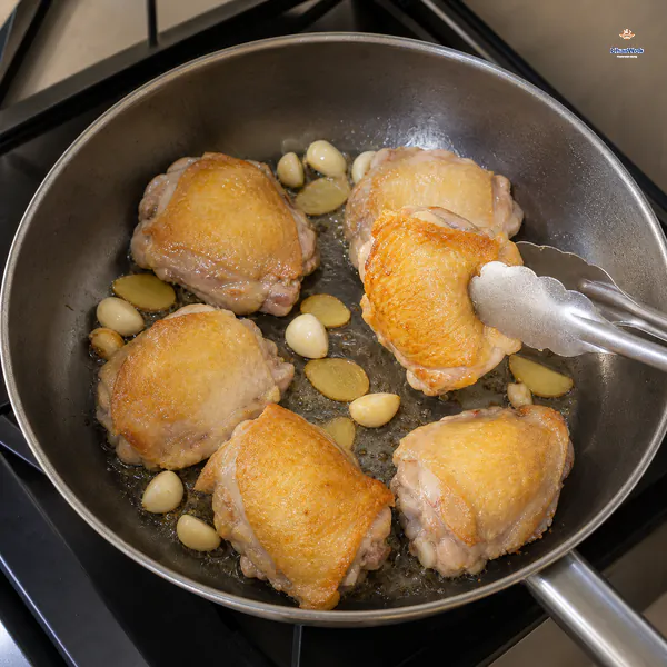 Six chicken thighs sear skin-side down with whole garlic cloves and ginger slices as tongs lift one deeply golden piece