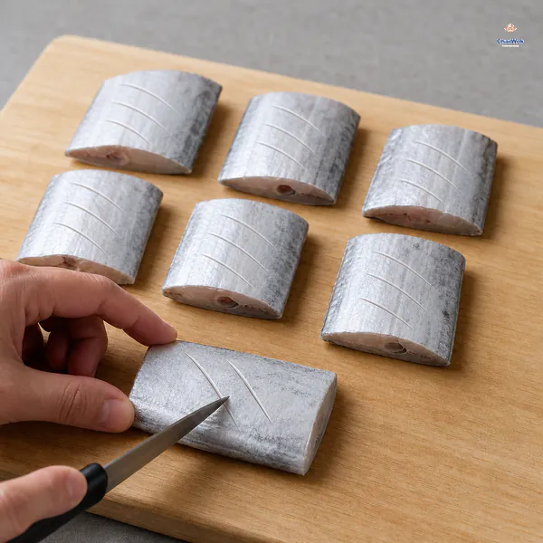 Seven equal belt-fish sections on a board as a knife makes the third shallow diagonal score in the foreground piece