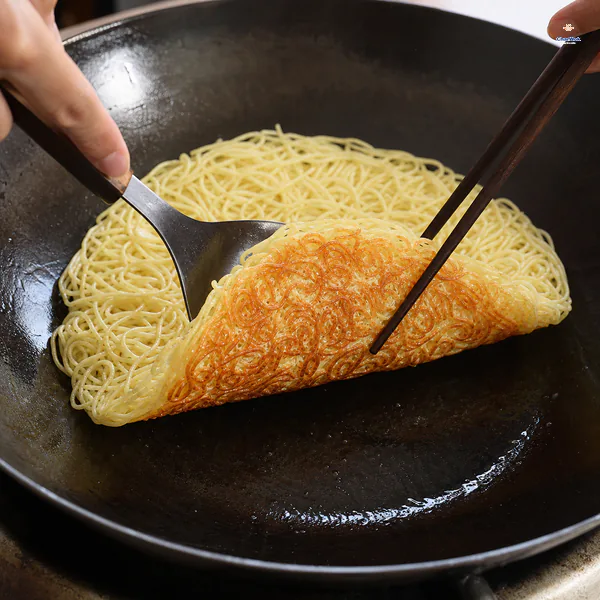 A spatula and chopsticks lift one edge of a thin noodle cake to reveal a continuous golden crisp underside in a carbon-steel pan