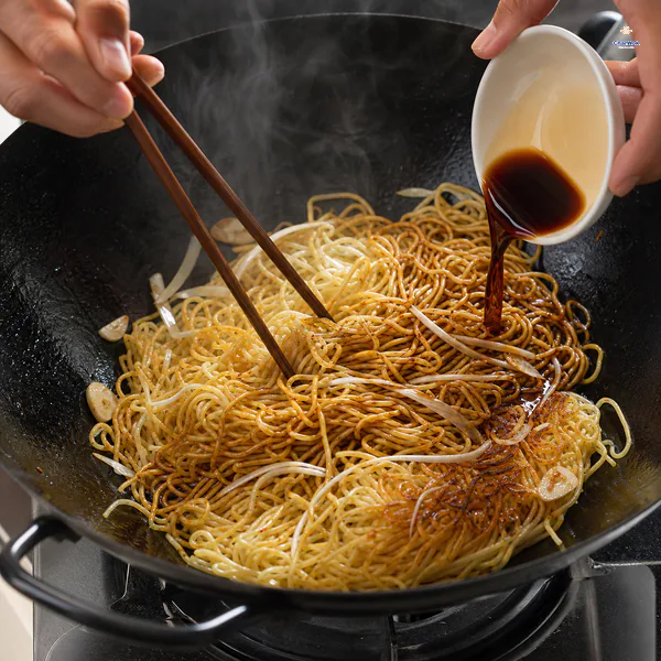A small dish pours soy sauce mixture along the hot wok edge as chopsticks toss partly golden and partly glazed thin noodles