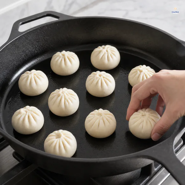 Raw pleated pork buns sit well spaced in a thin film of neutral oil as the last bun is placed in a cast-iron pan