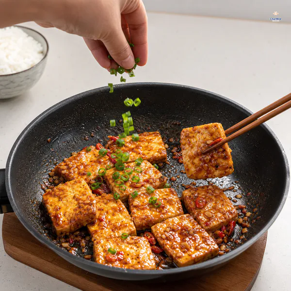 Scallion greens fall over glossy garlic-sauce tofu while chopsticks lift one square carrying a thin bright coating