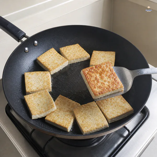 A thin spatula lifts one tofu square to reveal a complete deep-amber crust while the uncrowded pieces fry in a shallow oil film