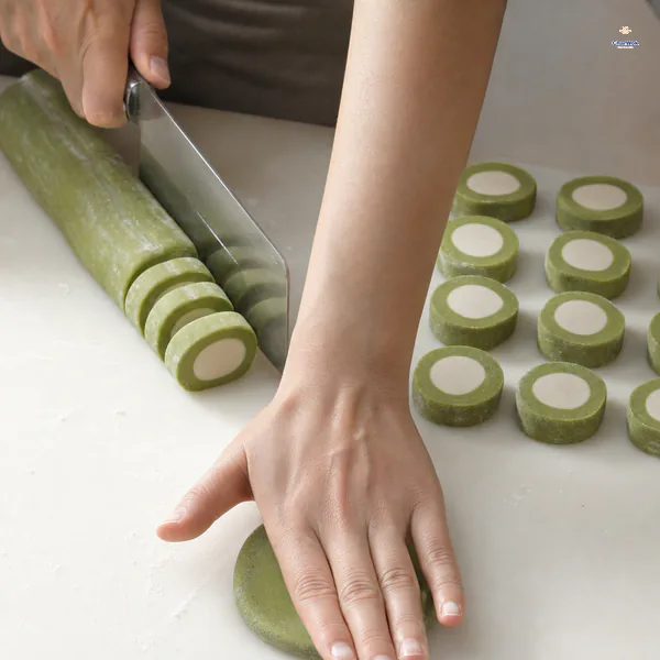 A green-wrapped white-core dough log is cut into even concentric slices while one slice is pressed flat with its white circle facing up