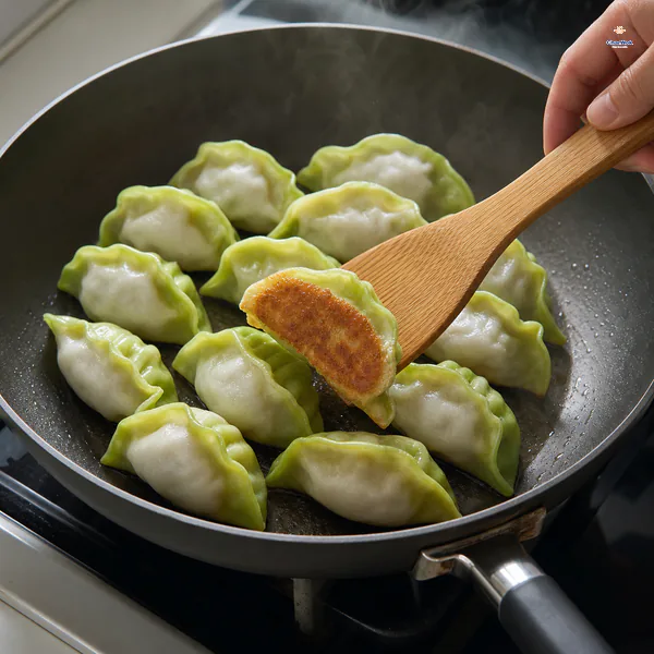 A wooden spatula lifts one green-and-white dumpling to reveal a deeply golden flat bottom while the remaining dumplings sear in a thin oil film