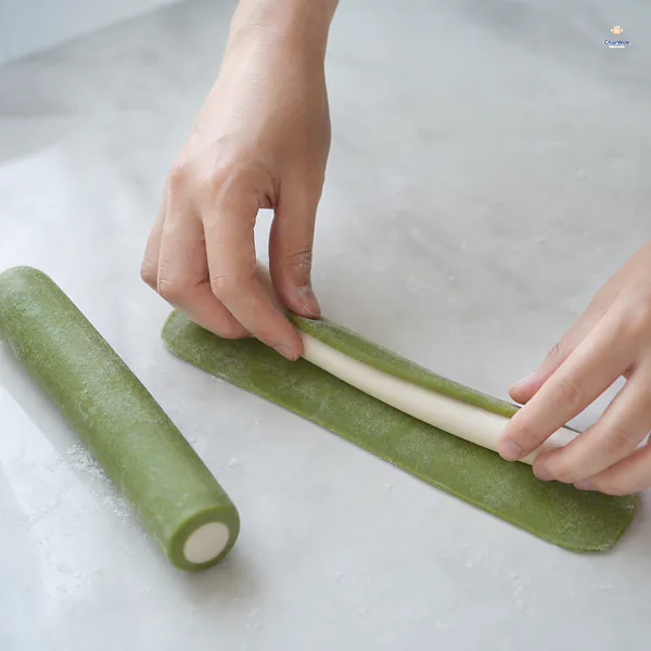 A thin spinach-green dough rectangle wraps around a white dough rope while a finished cylinder shows its green outer ring and white core