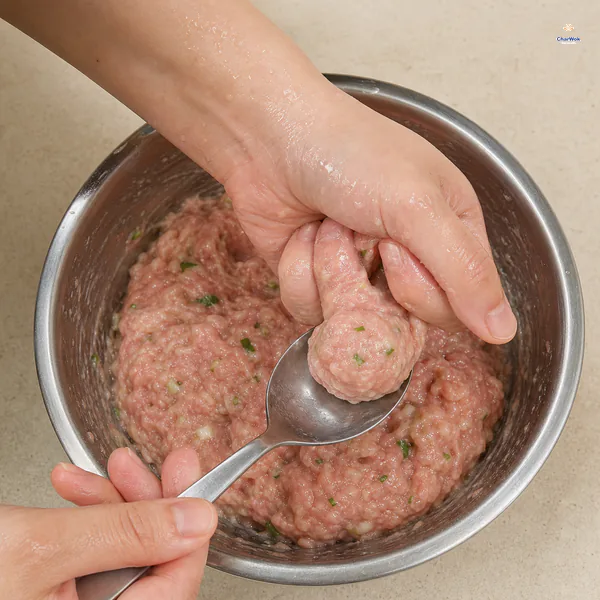 One damp hand squeezes a smooth raw pork ball through the opening between thumb and index finger while a spoon lifts it above the mixing bowl