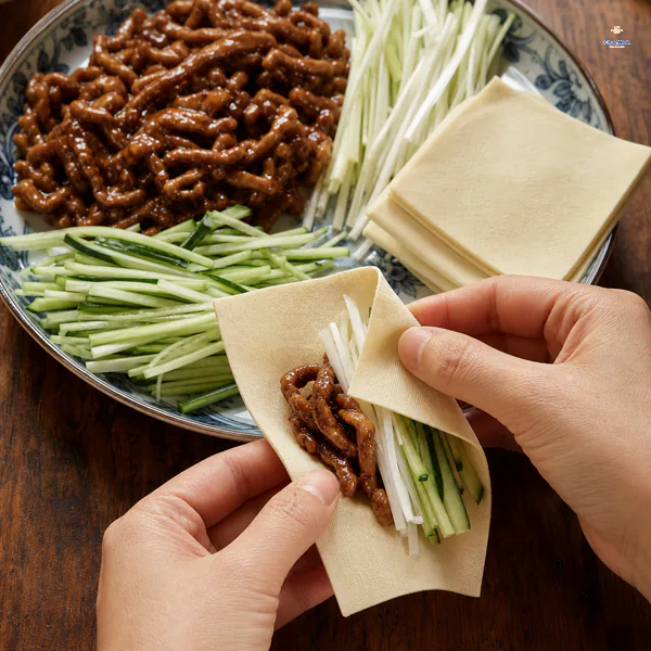 Hands fold a soft square tofu sheet around glossy pork slivers, white scallion and seeded cucumber matchsticks