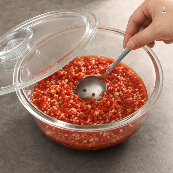 A clean spoon presses coarse chili solids beneath their own brine in a glass bowl with its lid visibly ajar