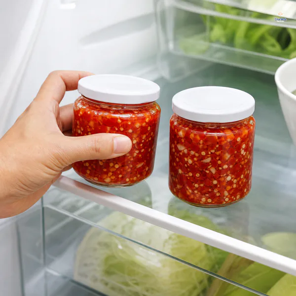 Two clean sealed jars of chunky red chili garlic sauce sit inside a refrigerator with solids beneath their own liquid