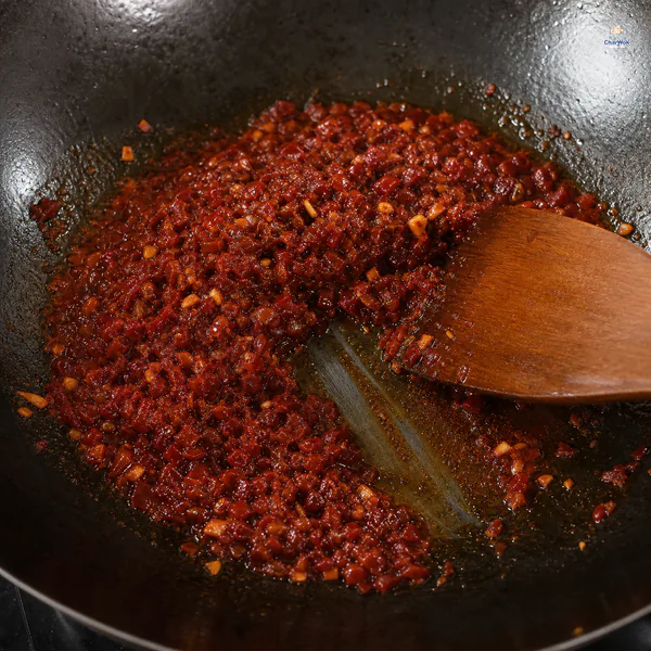 Deep brick-red chopped doubanjiang with tiny broad-bean pieces fries until a clear ruby oil halo forms and a spatula exposes the wok bottom