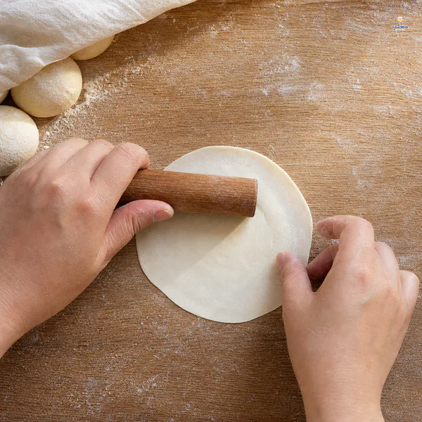 A small rolling pin thins the rim of a round dumpling wrapper while leaving its centre visibly thicker