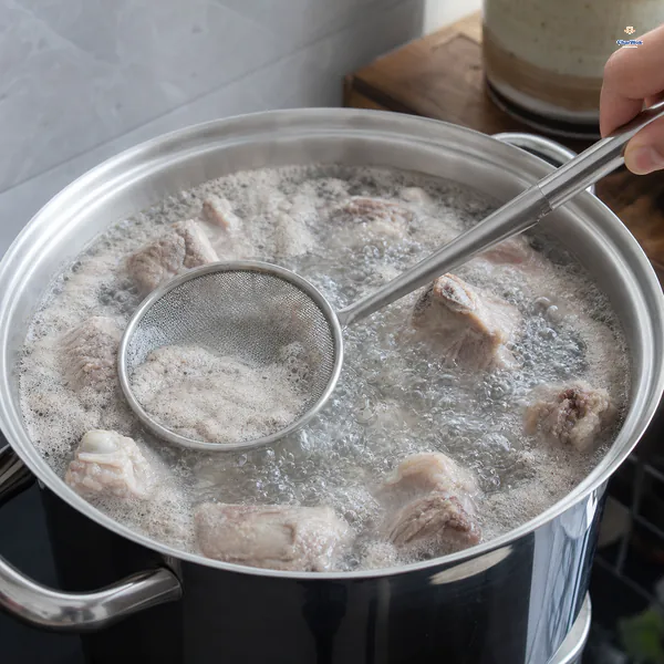 A fine-mesh ladle skims grey-white foam from pork ribs at a full boil in their cold-start blanching water