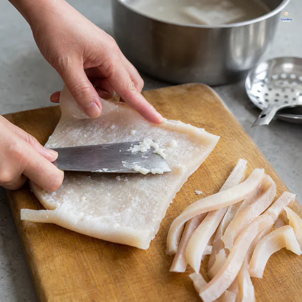 A knife scrapes the last white fat from warm blanched pork skin beside narrow cleaned strips