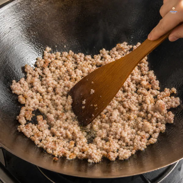 A wooden spatula breaks ground pork into small loose crumbles with rendered fat and a few lightly browned edges