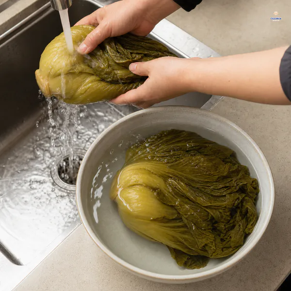 Whole yellow-green pickled mustard greens are rinsed under running water beside a bowl where another whole green soaks