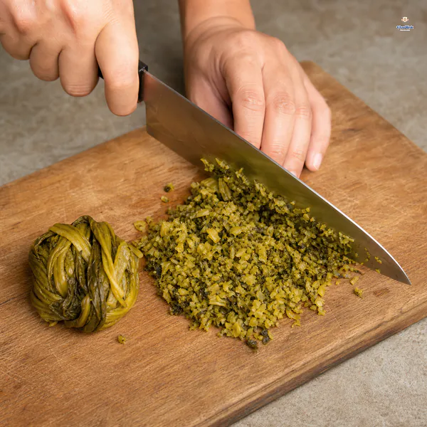 A squeezed-dry clump of pickled mustard green sits beside a knife chopping the rest into very fine pieces