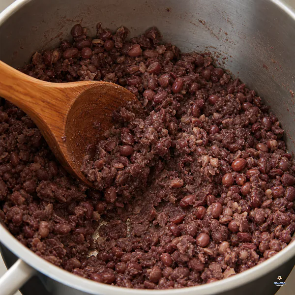 A broad wooden ladle presses well-drained adzuki beans into a dry-moist half-mash with many bean halves still intact
