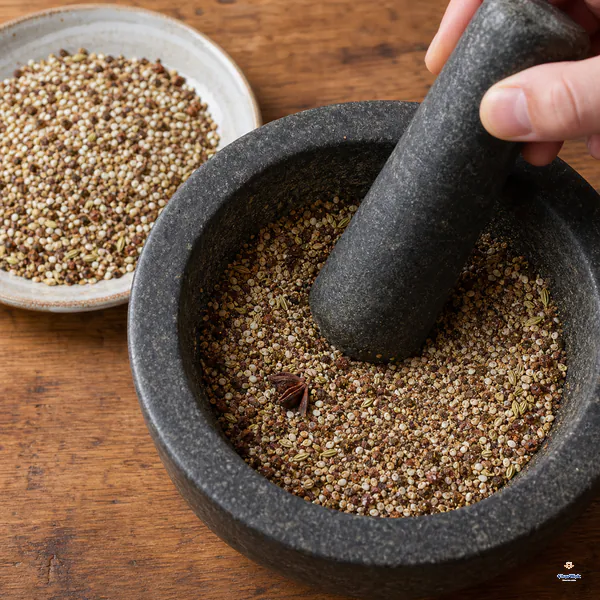 A fully cooled toasted spice-and-salt blend is being ground to a slightly coarse texture in a stone mortar beside the unground blend