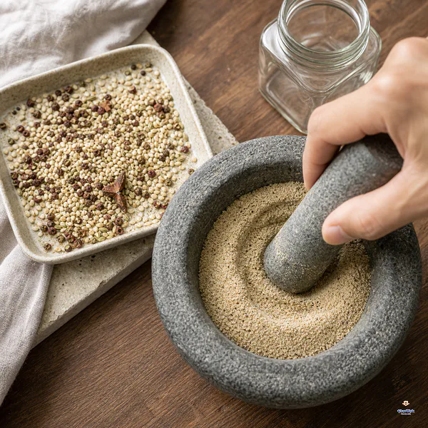 Completely cooled toasted spices and salt are ground into a fine dry beige pepper-salt powder in a stone mortar beside a plain jar