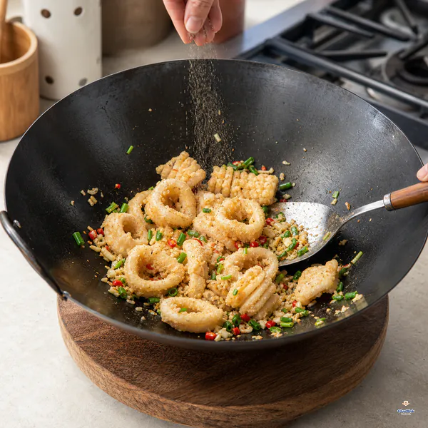 A wok rests off the burner on a wooden trivet while lightly golden crisp squid, scallions and aromatics receive a fine even veil of pepper-salt