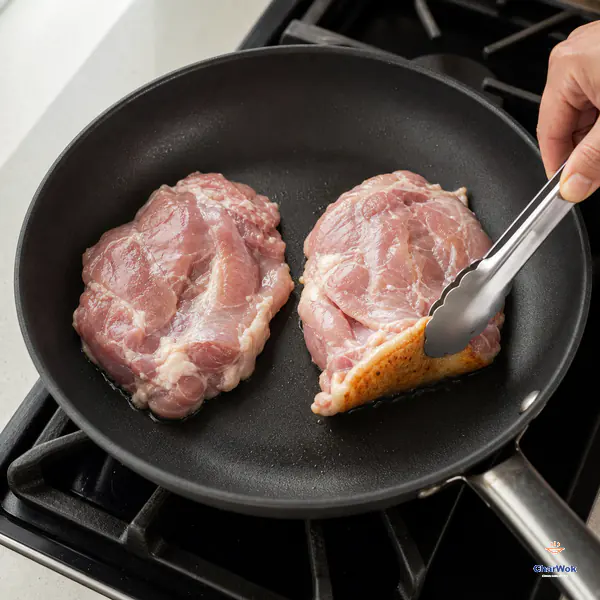 Two flattened boneless chicken thighs cook skin-side down in a dry skillet while one lifted corner shows golden skin and rendered fat