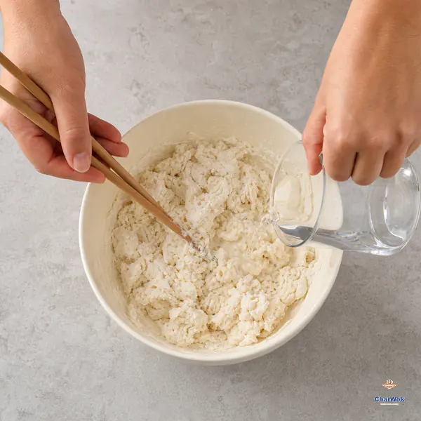 Lukewarm water pours into flour, yeast, baking powder and sugar while chopsticks form a damp shaggy mixture with no loose flour