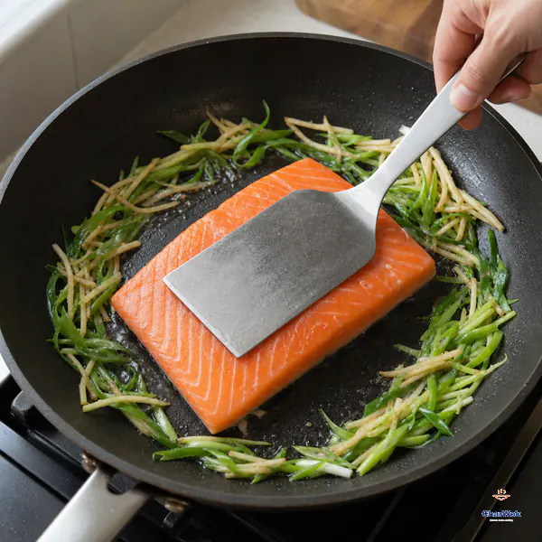 A wide spatula presses a salmon fillet flat with orange flesh facing up and its skin against the pan while scallion and ginger sit at the edge