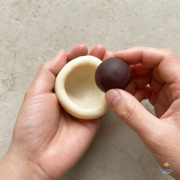 Two hands hold one glutinous-rice dough portion shaped into a deep thick-based bowl and one smooth red bean paste ball above its cavity
