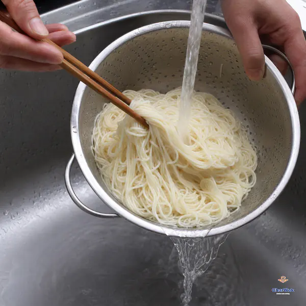 Cold running water rinses thin cooked noodles in a tilted metal colander while chopsticks lift and separate the strands