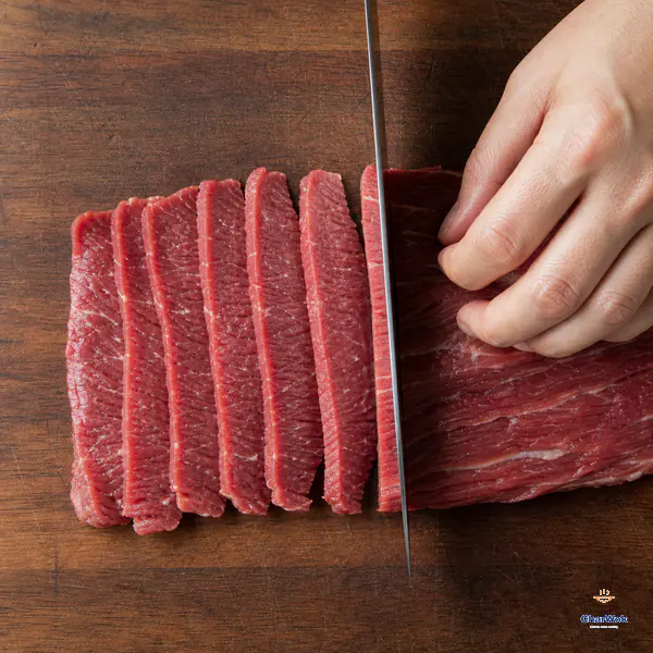 A chef's knife cuts raw beef perpendicular to the visible muscle fibres into thin slices about three millimetres thick