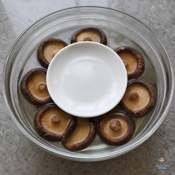Eight shiitake caps sit fully below cold clear water in a wide glass bowl under a small clean ceramic saucer