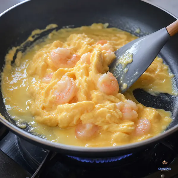 A silicone spatula slowly pushes shrimp and pale egg into large glossy folds while liquid egg flows back onto the low-heated pan