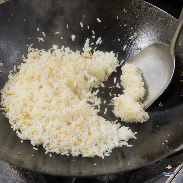 A metal wok spatula presses apart the final cold-rice clumps while most pale grains toss separately in a hot carbon-steel wok