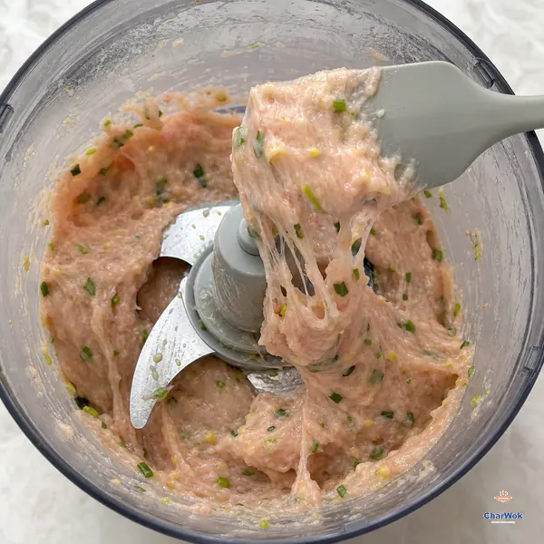 A spatula lifts pale shrimp paste from a stopped food processor and the cohesive mixture pulls back in several thick tacky strands