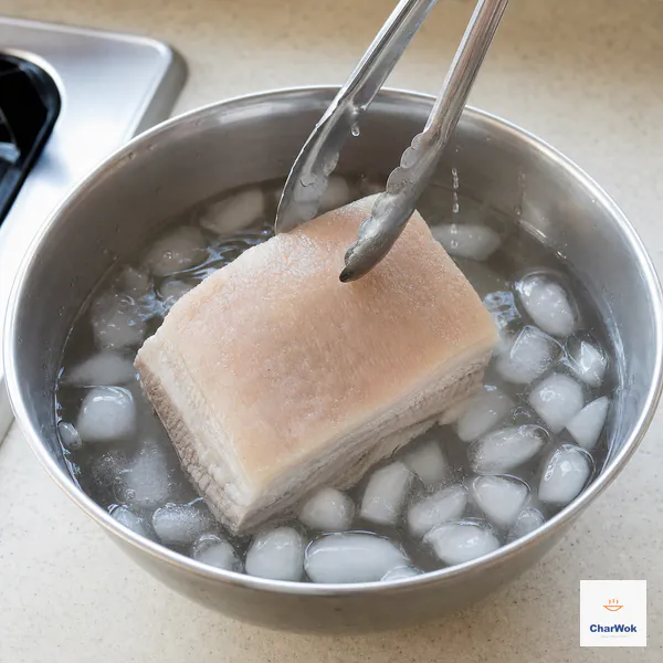 Metal tongs lower a fully cooked intact pork belly slab into a stainless-steel bowl crowded with clear ice cubes