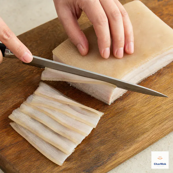 A long slicing knife cuts a chilled skin-on pork belly slab across its wide face beside flexible two-millimeter slices