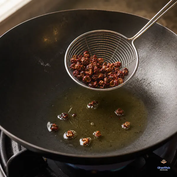 A small wire skimmer lifts fragrant whole red Sichuan pepper husks from clear pale-gold oil after a gentle low-heat infusion