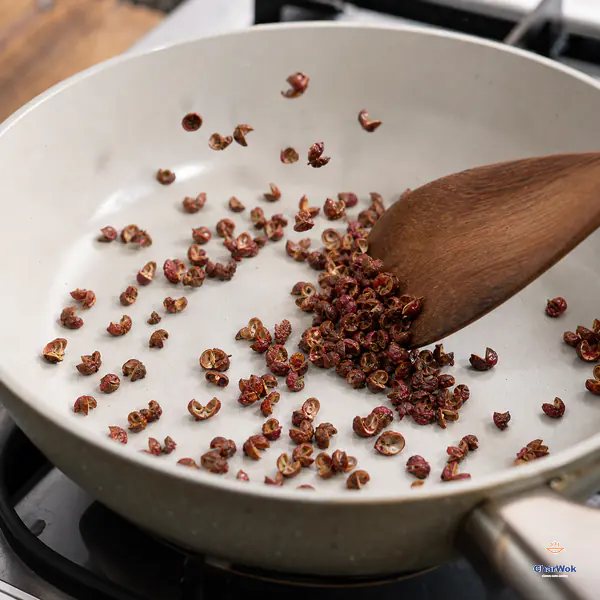 A dry wooden spatula moves an uncrowded layer of red Sichuan pepper husks in a completely dry pale skillet