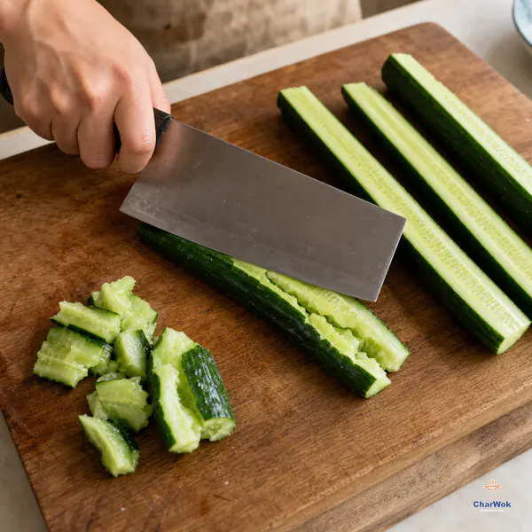 The flat side of a Chinese cleaver cracks one cucumber quarter into connected jagged ribbons beside firm rough-edged chunks