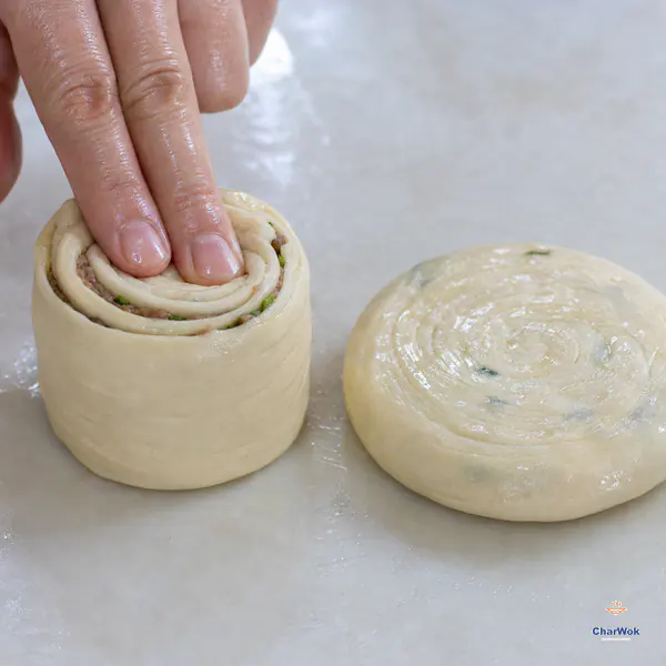 Two fingertips press straight down on an upright spiral dough cylinder beside an intact round uncooked disc with visible concentric layers